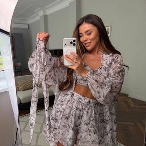 May include: A three-piece matching set featuring a light-colored crop top, high-waisted shorts, and a long-sleeved button-down shirt. The garments are made of a sheer, lightweight fabric decorated with a dark gray and black abstract floral print.