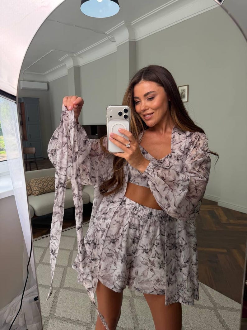 May include: A three-piece matching set featuring a light-colored crop top, high-waisted shorts, and a long-sleeved button-down shirt. The garments are made of a sheer, lightweight fabric decorated with a dark gray and black abstract floral print.