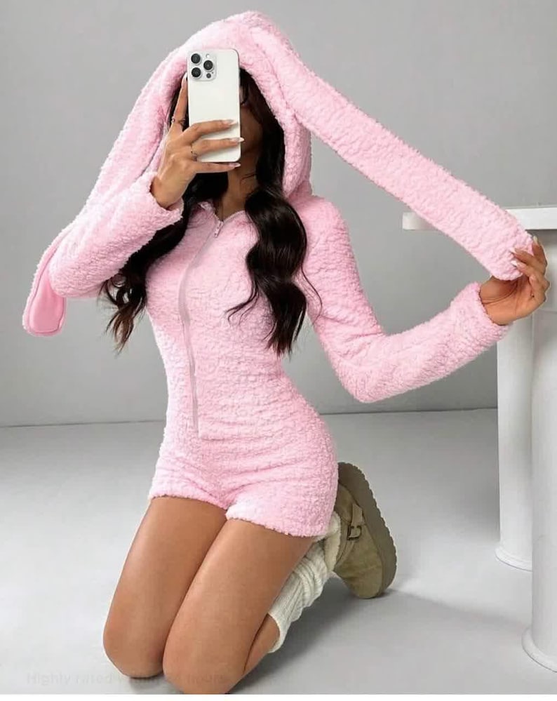 May include: A light pink, fuzzy romper with a hood featuring long bunny ears. The one-piece outfit has a zipper down the front and short shorts. The person in the image is wearing white socks and tan boots.