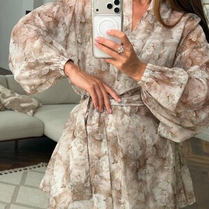 May include: A beige and white floral print wrap dress featuring long, voluminous bishop sleeves with elasticized cuffs, a V-neckline, and a matching fabric tie belt at the waist. The garment is crafted from a lightweight, semi-sheer fabric.