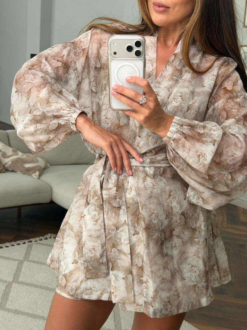 May include: A beige and white floral print wrap dress featuring long, voluminous bishop sleeves with elasticized cuffs, a V-neckline, and a matching fabric tie belt at the waist. The garment is crafted from a lightweight, semi-sheer fabric.