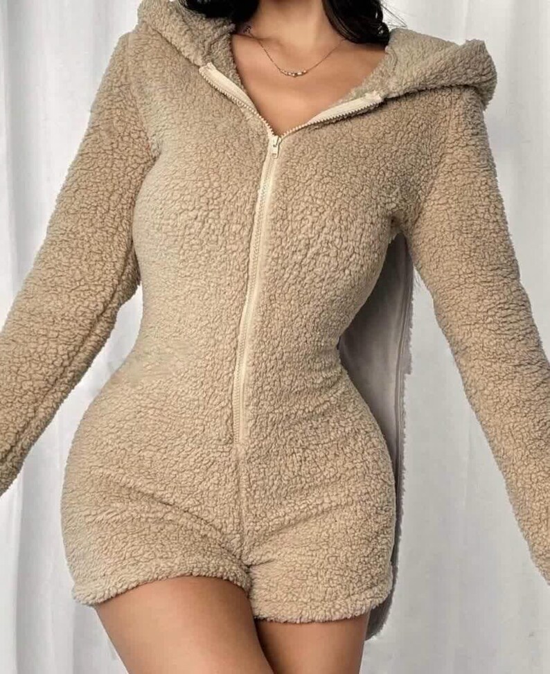 May include: A beige, hooded romper with a full-length zipper and short shorts. The romper is made of a soft, textured material. The long sleeves and hood provide warmth, while the shorts offer a casual look. The outfit is designed for comfort and style.