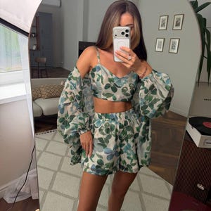 May include: A two-piece set consisting of a cropped camisole top with thin straps and matching high-waisted shorts, both featuring a green and blue botanical leaf print on a light background, paired with a long-sleeved, sheer, matching cover-up.