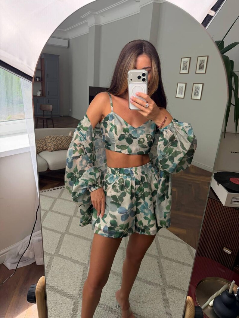 May include: A two-piece set consisting of a cropped camisole top with thin straps and matching high-waisted shorts, both featuring a green and blue botanical leaf print on a light background, paired with a long-sleeved, sheer, matching cover-up.