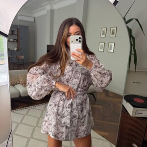 May include: A long-sleeved mini dress featuring a light gray and black floral pattern. The dress has a high neckline, voluminous sleeves with gathered cuffs, and a matching fabric tie belt at the waist to create a cinched silhouette.