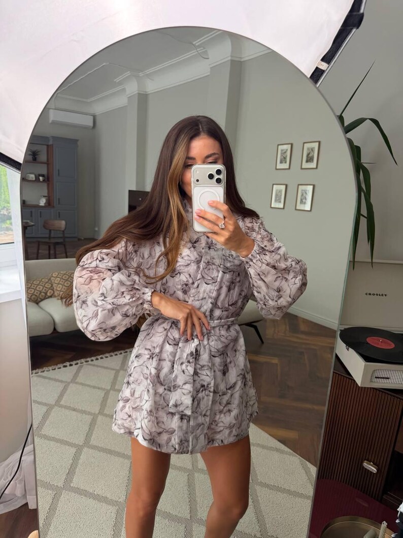 May include: A long-sleeved mini dress featuring a light gray and black floral pattern. The dress has a high neckline, voluminous sleeves with gathered cuffs, and a matching fabric tie belt at the waist to create a cinched silhouette.