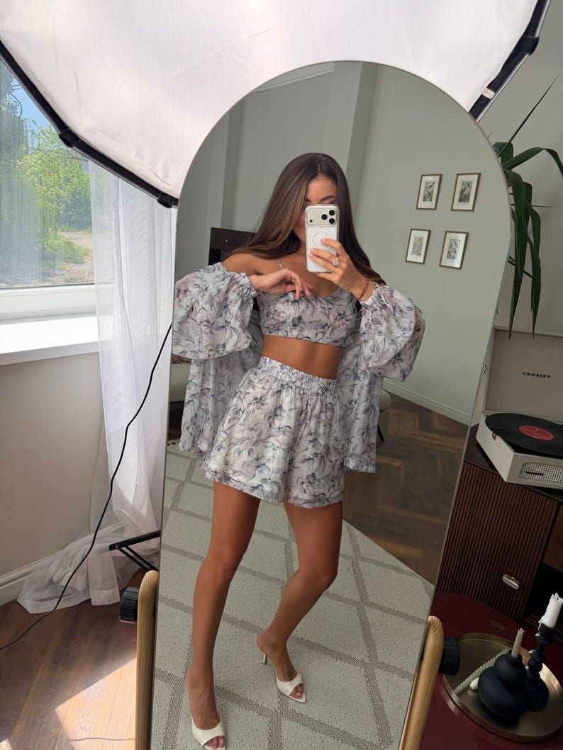 May include: A two-piece set consisting of a cropped camisole top and high-waisted shorts, paired with a matching long-sleeved button-down shirt. The garments feature a light-colored fabric with a dark, abstract, marbled floral print pattern.