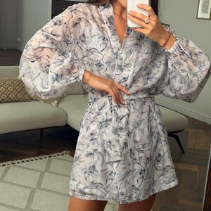 May include: A short, long-sleeved dress made of a sheer, lightweight fabric featuring a blue and grey floral pattern. The dress has a V-neckline, gathered cuffs at the wrists, and a matching fabric tie belt that cinches at the waist.