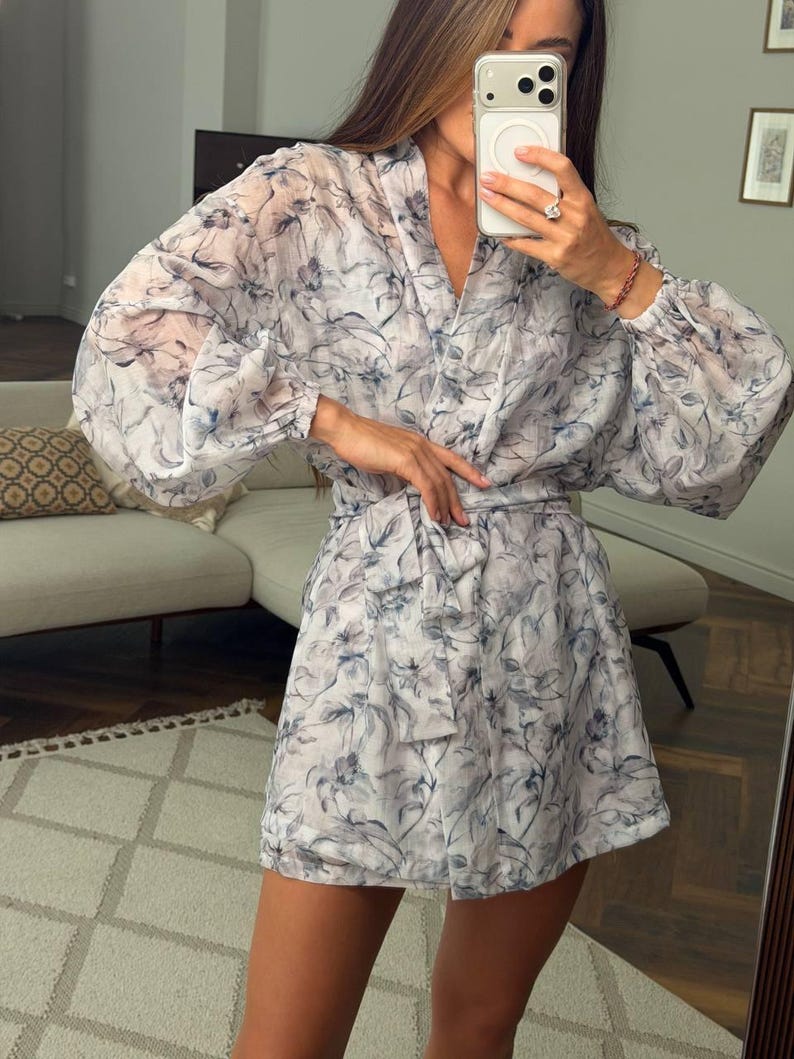 May include: A short, long-sleeved dress made of a sheer, lightweight fabric featuring a blue and grey floral pattern. The dress has a V-neckline, gathered cuffs at the wrists, and a matching fabric tie belt that cinches at the waist.