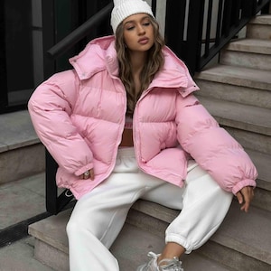Könnte beinhalten: Eine hellrosa Pufferjacke mit hohem Kragen und einer weißen Mütze. Die Person trägt eine weiße Jogginghose und graue Turnschuhe. Die Jacke scheint aus einem gesteppten Material zu sein.