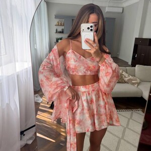 May include: A three-piece set featuring a cropped camisole top, a pair of high-waisted shorts, and a long-sleeved open cardigan. Each piece is made from a lightweight fabric with a soft pink and white tie-dye pattern throughout the entire set.