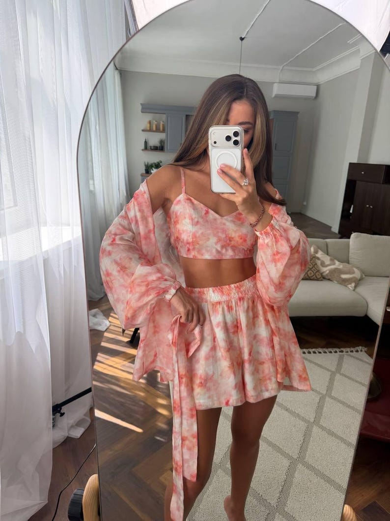May include: A three-piece set featuring a cropped camisole top, a pair of high-waisted shorts, and a long-sleeved open cardigan. Each piece is made from a lightweight fabric with a soft pink and white tie-dye pattern throughout the entire set.