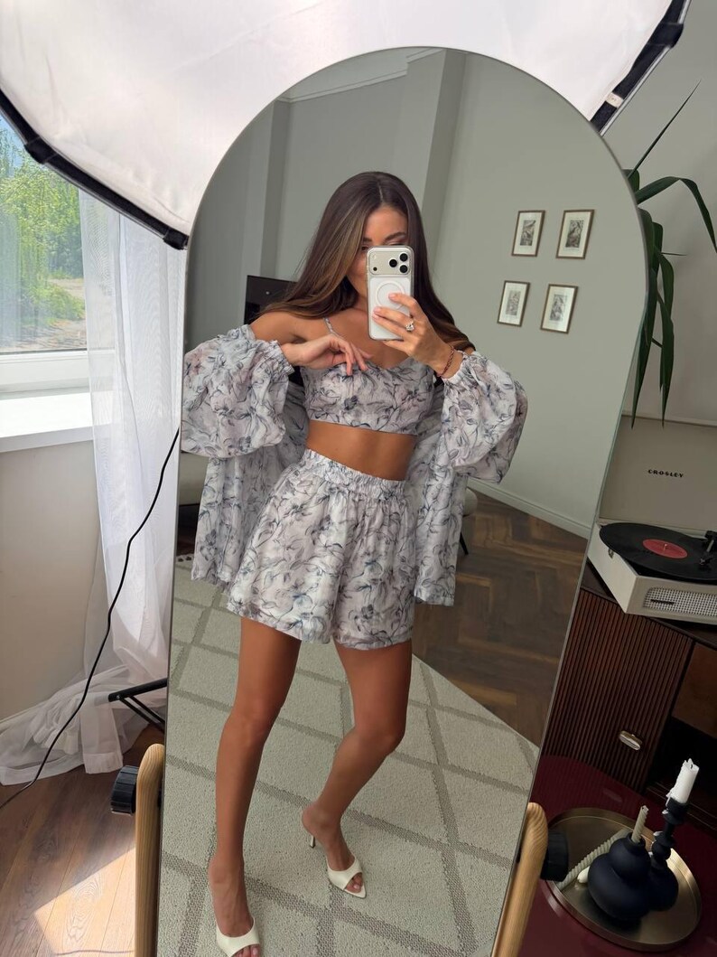May include: A three-piece matching set featuring a cropped camisole top, high-waisted shorts, and a long-sleeved button-down shirt. The garments are made of a lightweight, semi-sheer fabric with a grey and white abstract floral pattern.