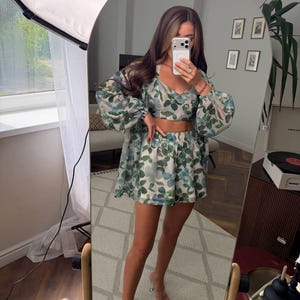 May include: A three-piece matching set featuring a cropped camisole top, high-waisted shorts, and a long-sleeved sheer button-down shirt. The garments are made of a lightweight fabric with a botanical print of green and blue leaves on a white base.