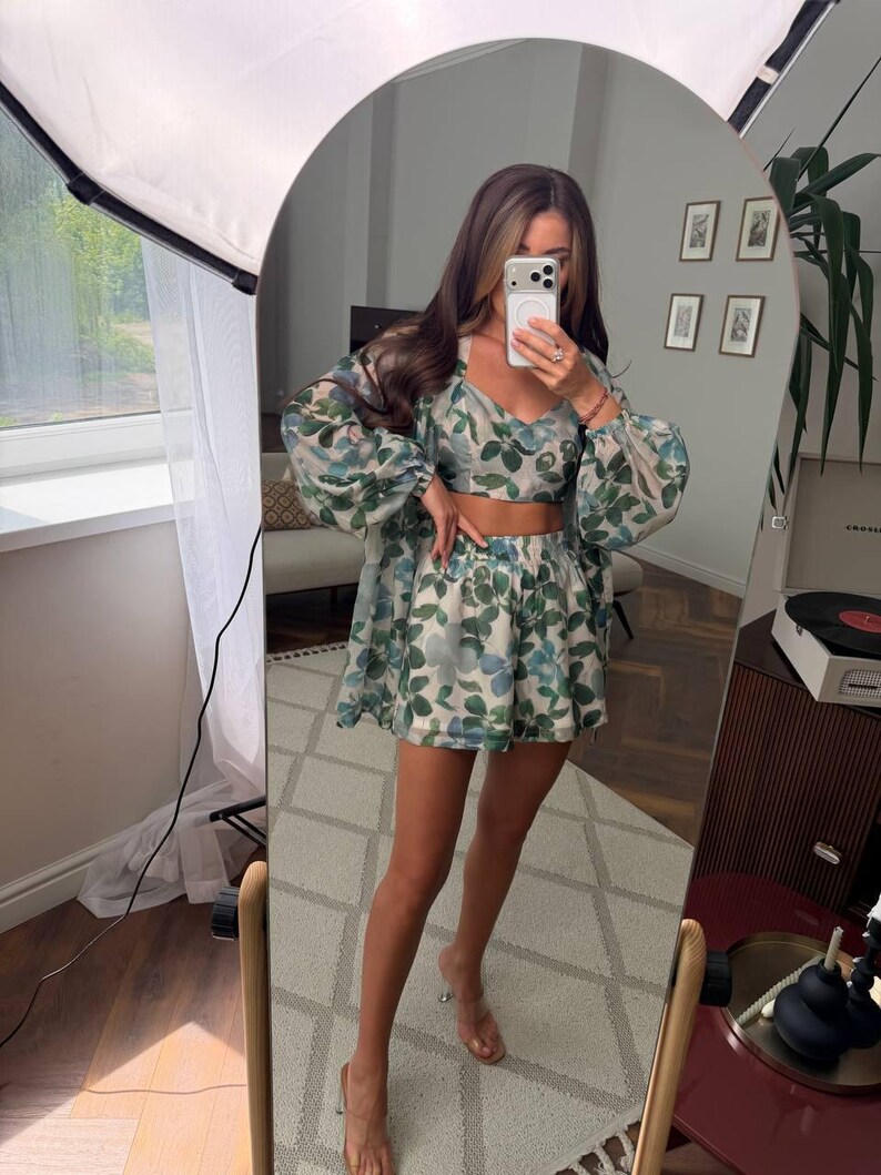May include: A three-piece matching set featuring a cropped camisole top, high-waisted shorts, and a long-sleeved sheer button-down shirt. The garments are made of a lightweight fabric with a botanical print of green and blue leaves on a white base.