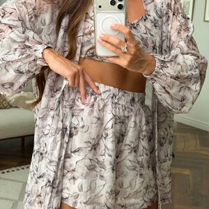May include: A three-piece set consisting of a long-sleeved, open-front duster cardigan, a cropped tank top, and high-waisted shorts. The garments are made of a lightweight, sheer fabric featuring a monochromatic gray and white floral print.