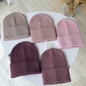 Könnte beinhalten: Fünf gerippte Strickmützen in verschiedenen Rosa- und Lilatönen liegen auf einer weißen Oberfläche. Die Mützen sind am Rand umgeschlagen und haben ein schlichtes, klassisches Design. Die Farben reichen von hellem Mauve bis zu tiefem Pflaume.