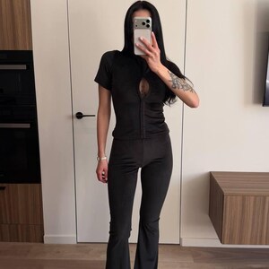 Puede incluir: Conjunto de dos piezas de terciopelo negro. La parte superior tiene cremallera, mangas cortas y un detalle de ojo de cerradura. Los pantalones son acampanados. El conjunto se usa con zapatos negros. Ideal para estar en casa.