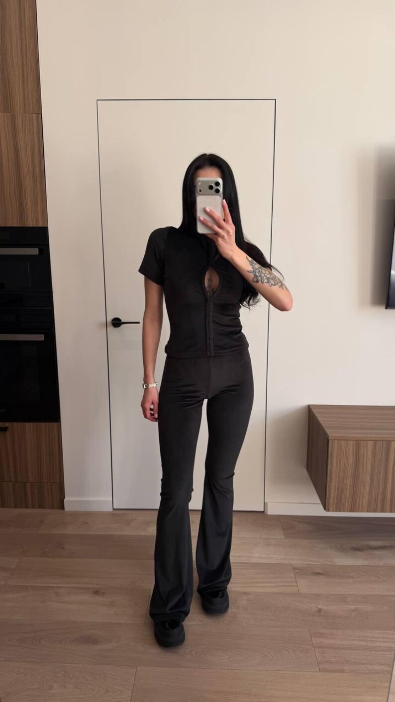 Puede incluir: Conjunto de dos piezas de terciopelo negro. La parte superior tiene cremallera, mangas cortas y un detalle de ojo de cerradura. Los pantalones son acampanados. El conjunto se usa con zapatos negros. Ideal para estar en casa.