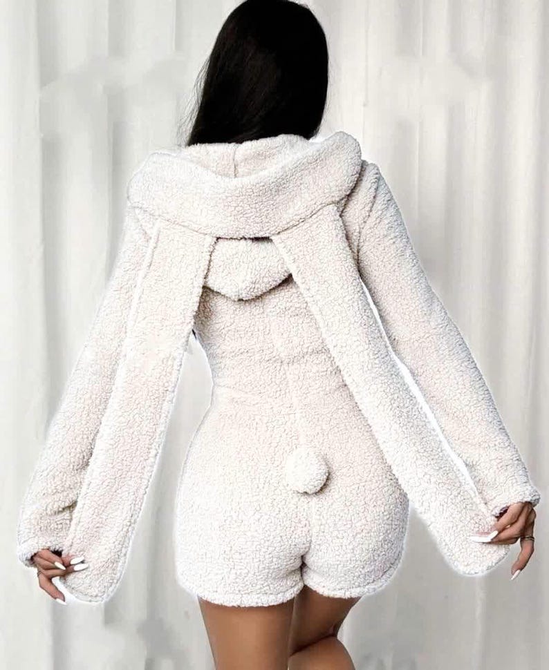 May include: A cream-colored, hooded romper with long sleeves and shorts. The romper features long, floppy bunny ears attached to the hood and a small, fluffy tail on the back. The fabric has a soft, textured appearance.