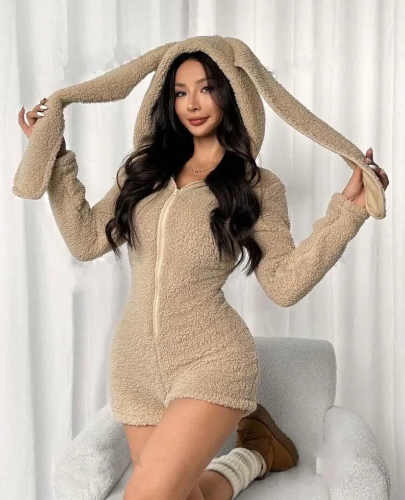 May include: A beige, hooded onesie with long sleeves and shorts, featuring a bunny ear design on the hood. The onesie has a full-length zipper and is made of a soft, textured material. The model is wearing brown boots and white socks.