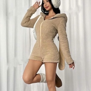 May include: A tan, hooded romper with a zip-up front and shorts. The romper has long sleeves and a fuzzy texture. The outfit includes a pair of bunny ears and white earmuffs. The model is also wearing white socks and brown boots.