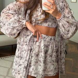 May include: A three-piece set consisting of a long-sleeved, button-down sheer shirt, a cropped top, and elastic-waist shorts. The garments feature a light-colored base with a dark, abstract floral print throughout the fabric.