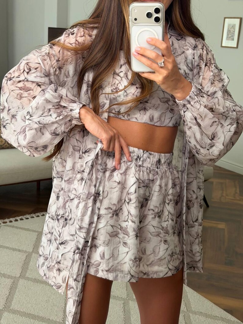 May include: A three-piece set consisting of a long-sleeved, button-down sheer shirt, a cropped top, and elastic-waist shorts. The garments feature a light-colored base with a dark, abstract floral print throughout the fabric.