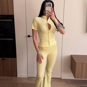 Puede incluir: Un ch&aacute;ndal de terciopelo amarillo claro, con una parte superior con cremallera de manga corta y pantalones acampanados. El conjunto es de color liso y parece estar hecho de un material suave y c&oacute;modo. La persona lleva zapatillas blancas.