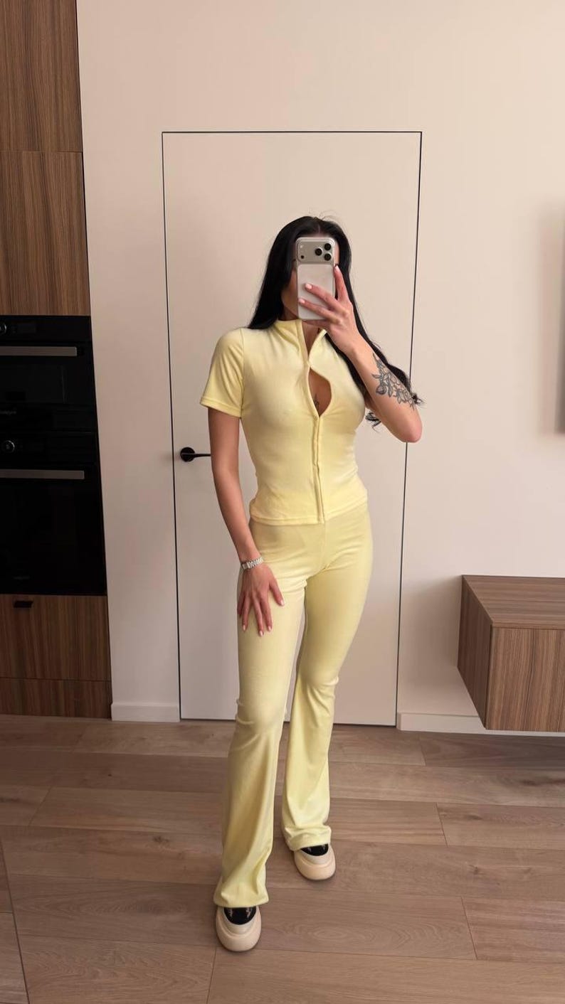 Puede incluir: Un ch&aacute;ndal de terciopelo amarillo claro, con una parte superior con cremallera de manga corta y pantalones acampanados. El conjunto es de color liso y parece estar hecho de un material suave y c&oacute;modo. La persona lleva zapatillas blancas.