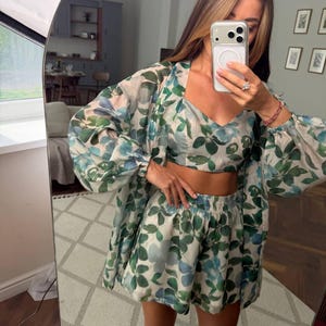 May include: A three-piece matching set featuring a cropped camisole top, high-waisted shorts with an elastic waistband, and a long-sleeved button-down shirt. The garments are made of a sheer, lightweight fabric with a green and blue leaf print.
