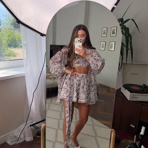 May include: A matching two-piece set consisting of a cropped top and high-waisted shorts, paired with a long-sleeved, button-down shirt. The garments feature a light-colored fabric with a dark, intricate floral or botanical print pattern throughout.