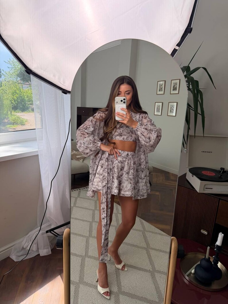 May include: A matching two-piece set consisting of a cropped top and high-waisted shorts, paired with a long-sleeved, button-down shirt. The garments feature a light-colored fabric with a dark, intricate floral or botanical print pattern throughout.