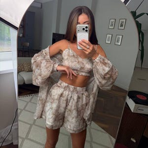 May include: A three-piece matching set featuring a cropped camisole top, high-waisted shorts, and a long-sleeved button-down shirt. The garments are made of a sheer, lightweight fabric with an all-over beige and brown floral pattern.