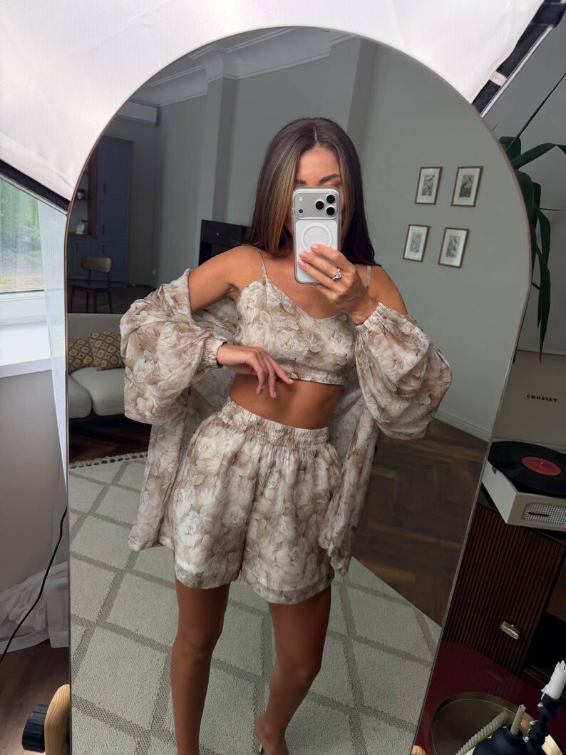 May include: A three-piece matching set featuring a cropped camisole top, high-waisted shorts, and a long-sleeved button-down shirt. The garments are made of a sheer, lightweight fabric with an all-over beige and brown floral pattern.