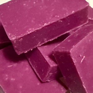 May include: Close-up of several rectangular bars of handmade soap. The soap is a deep magenta color, with a slightly textured surface. The bars are stacked and overlapping, creating a visually appealing arrangement.