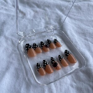 May include: A set of almond-shaped false nails in a clear plastic case. Each nail features a nude base with a black polka dot design on the tip. One nail has a small black floral design.