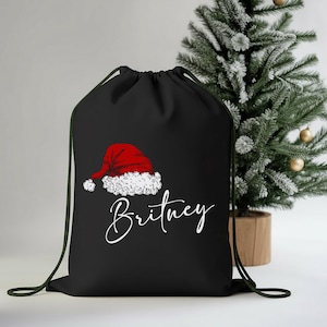 May include: Black drawstring bag featuring a red Santa hat graphic with white trim and the name "Britney" in a cursive font. A small Christmas tree with gold ornaments is in the background.