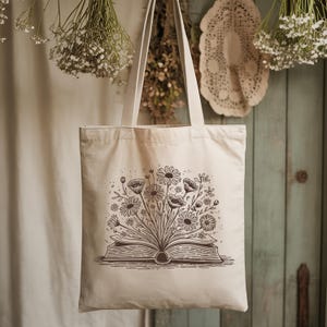 May include: A cream-colored tote bag with a floral design. The graphic depicts an open book with a bouquet of flowers bursting from its pages. The bag has long handles for carrying and is hanging against a rustic wooden background.