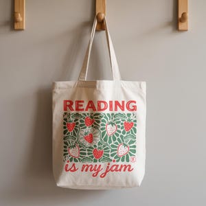 May include: A cream-colored canvas tote bag featuring a red and green graphic print of strawberries and foliage. The text READING is printed in red at the top, and the text is my jam is printed in red at the bottom of the strawberry design.