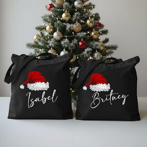 May include: Two black tote bags with a red Santa hat design and white text. One bag says "Isabel" and the other says "Britney". A Christmas tree with ornaments is in the background.