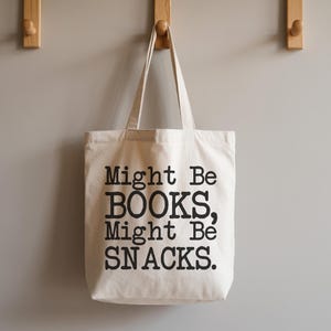 May include: A cream-colored tote bag with black text that reads "Might Be BOOKS, Might Be SNACKS." The bag hangs from a wooden hook on a gray wall. The bag has two handles.