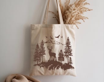May include: Beige tote bag with a brown forest scene featuring a deer, trees, mountains, and birds. The bag is hanging on a wooden hook. A tan blanket is draped below the bag.