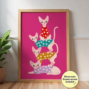 Op de afbeelding: Een ingelijste print met vier Sphynx-katten die op elkaar zijn gestapeld en elk een trui met stippen dragen in rood, teal, geel en paars. De achtergrond is felroze. De print wordt weergegeven op een houten vloer met een potplant.