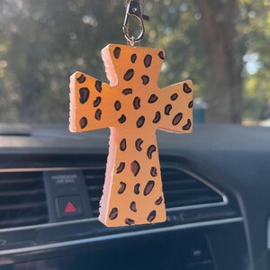 May include: A cross-shaped car charm with a leopard print design. The charm is light orange with dark brown spots and hangs from a black beaded chain. The charm is hanging in front of a car's dashboard.