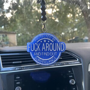 May include: Blue car air freshener with the text "PROTECTED BY FUCK AROUND AND FIND OUT SURVEILLANCE". The air freshener is round with a white border and is hanging from a black beaded chain.