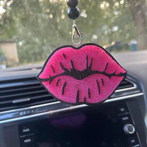 May include: A pink, lip-shaped car air freshener with a black outline and glitter accents. The air freshener hangs from a black beaded string and a silver clasp. The background shows a car interior and a blurred outdoor scene.