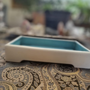 May include: A white ceramic mug with a black handle and the text "But First, Coffee" in a bold, black font. The mug is set against a blurred background, suggesting a cozy setting.