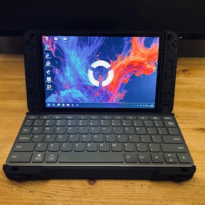 Puede incluir: Un ordenador portátil negro con un teclado gris y una pantalla que muestra un diseño abstracto colorido con un círculo blanco. El dispositivo está abierto y sobre una superficie de madera.