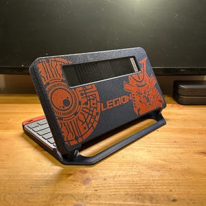 以下が含まれることがあります： Lenovo Legionハンドヘルドゲーム機用のダークブルーのテクスチャ付き保護ケース。ケースには、回路パターンと侍の兜の目立つオレンジ色のグラフィックデザインが施されており、中央にLEGIONという文字が印刷されています。